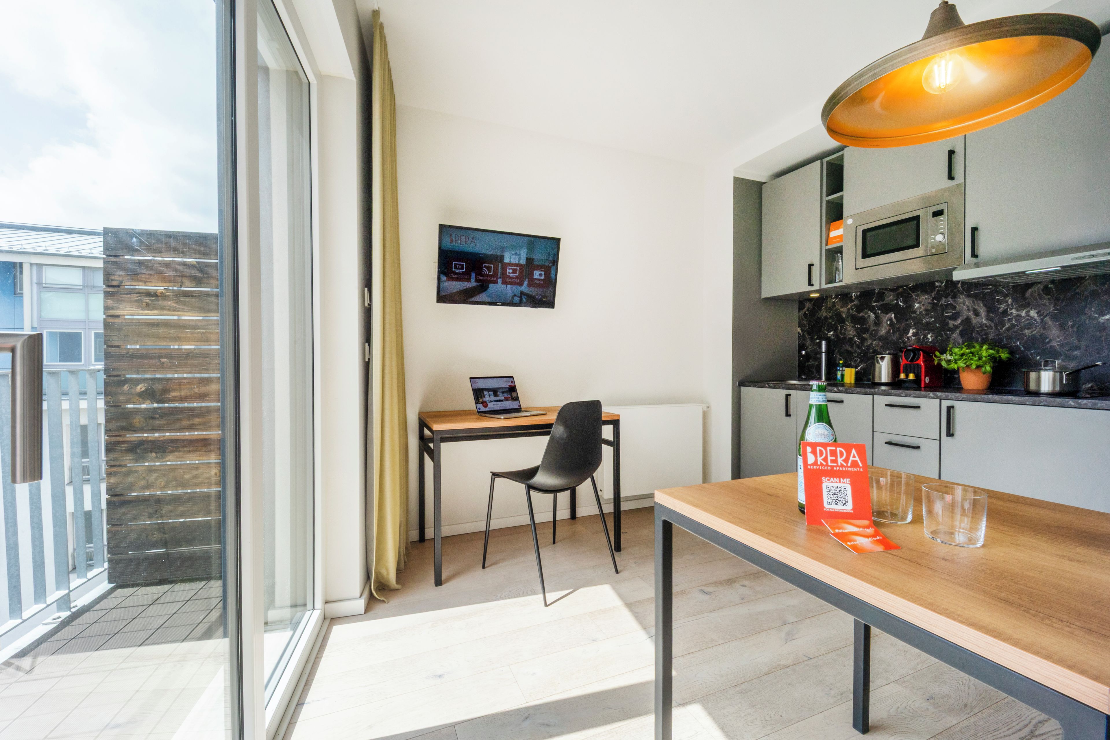 A modern kitchen with a dining area, a TV mounted on the wall, and a balcony with a view.