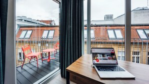 Una habitación con una laptop sobre una mesa de madera, una ventana con cortinas y vistas a un paisaje urbano con edificios y un balcón con sillas rojas.
