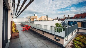 Terraza en la azotea con una silla y mesa rojas, baranda de metal y vista de edificios al fondo.