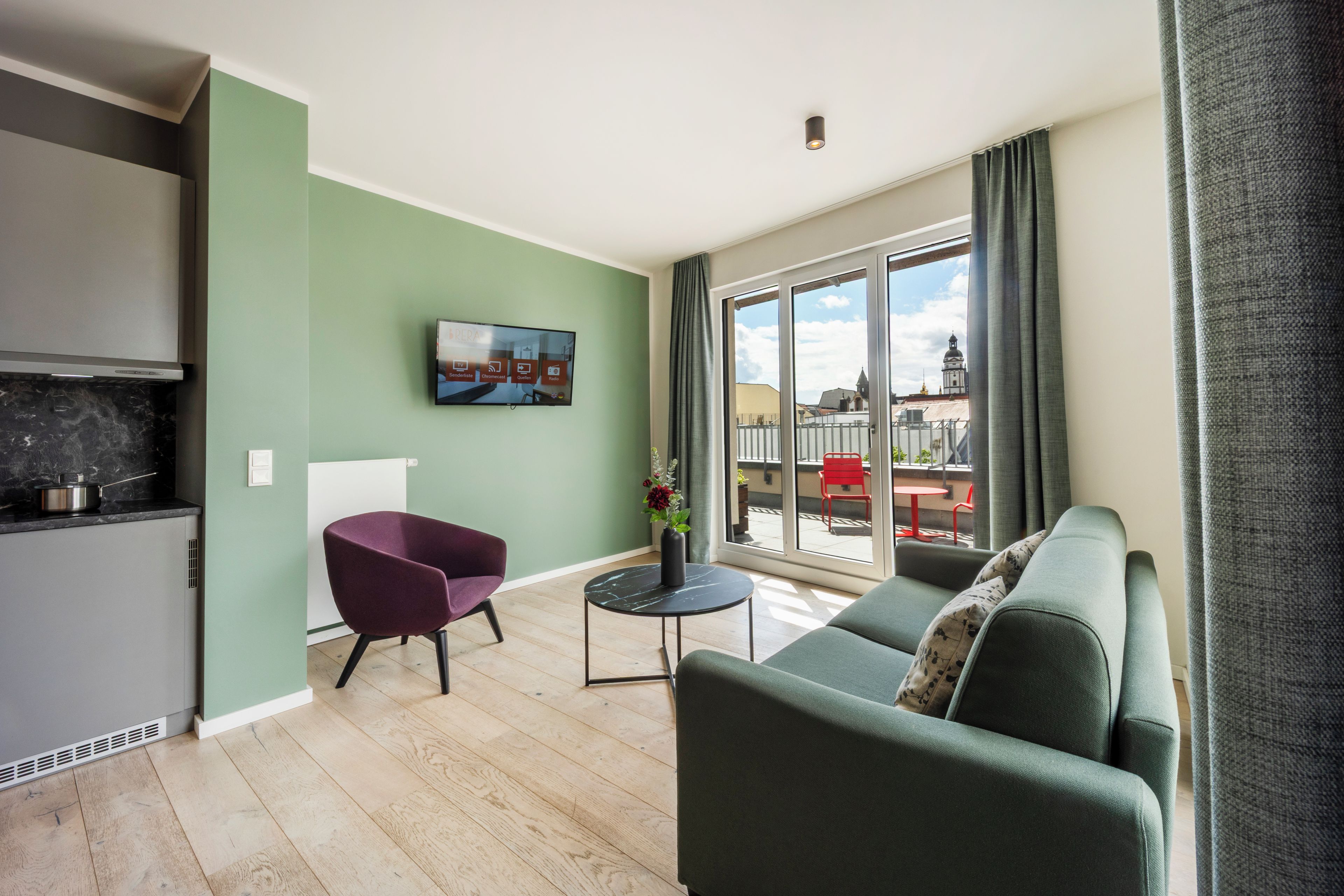 Een moderne woonkamer met een bank, een fauteuil, een aan de muur bevestigde televisie en uitzicht buiten door grote ramen.