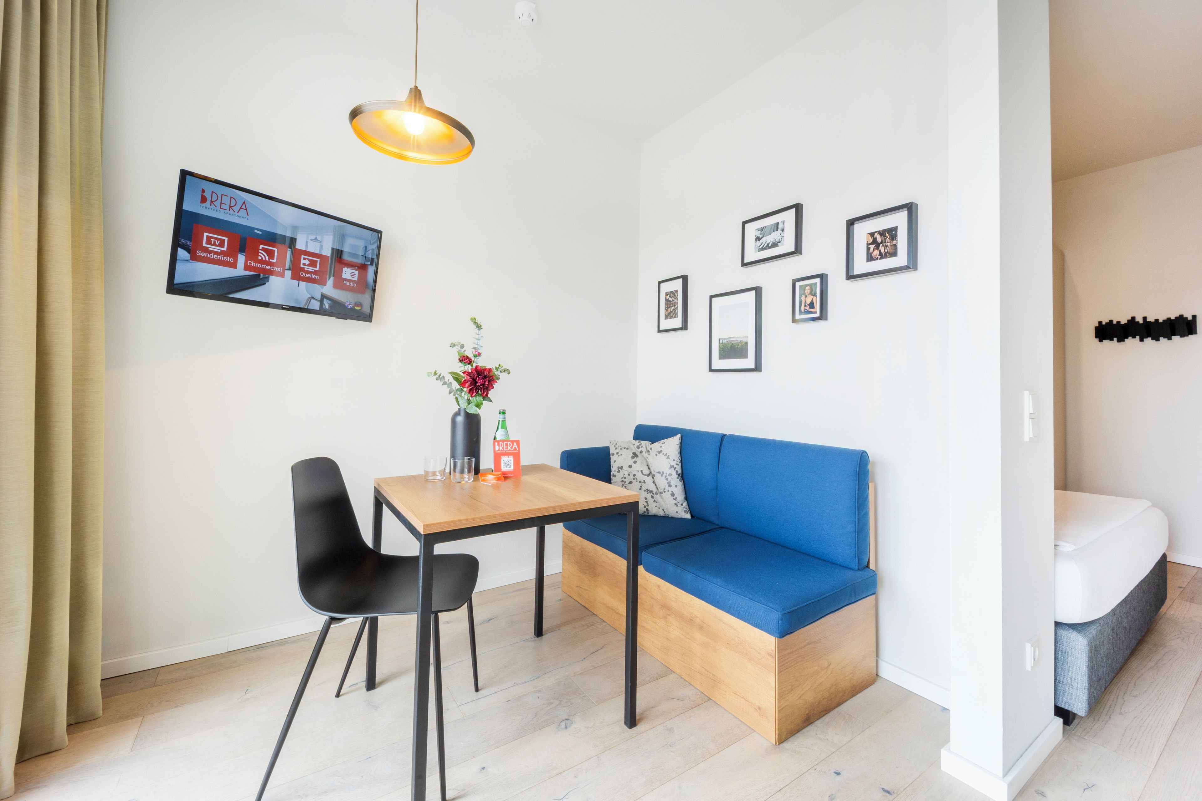 A modern living room with a blue sofa, a wooden table with two chairs, a pendant light, and a flat-screen TV mounted on the wall.