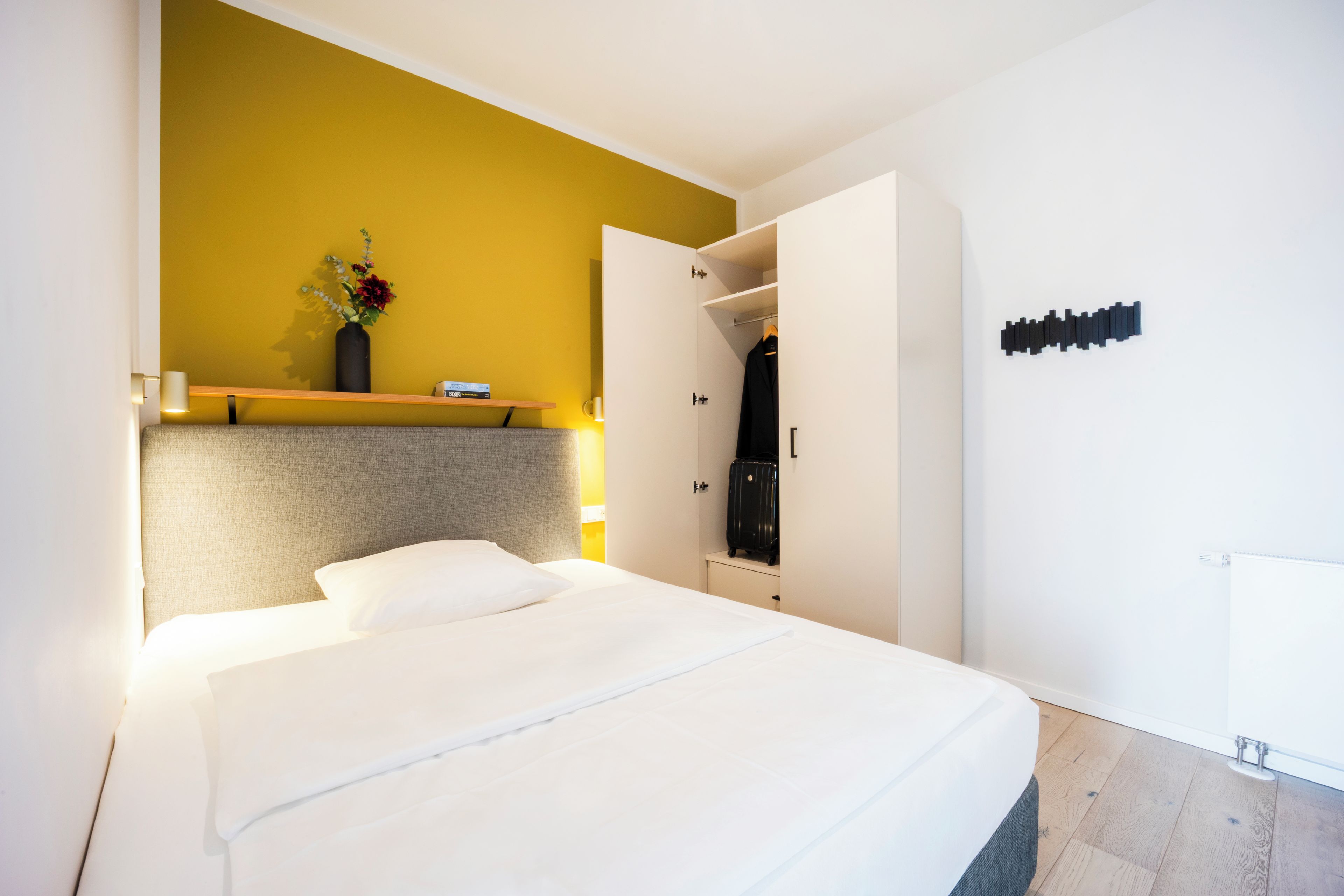 Una habitación de hotel con cama, mesita de noche, un jarrón con flores, armario y una lámpara de pared.