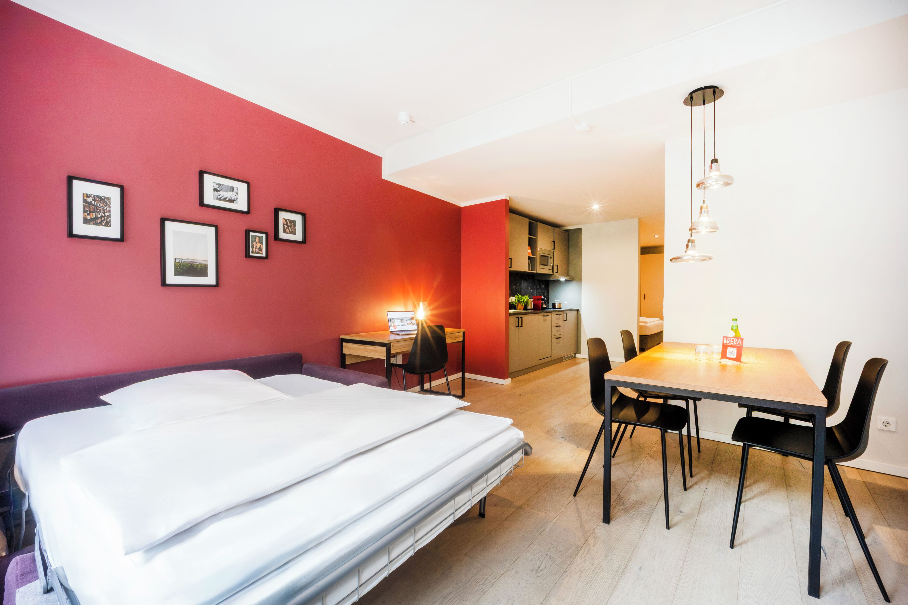 A hotel room with a bed, a desk, a dining table with chairs, and a kitchenette in the background.
