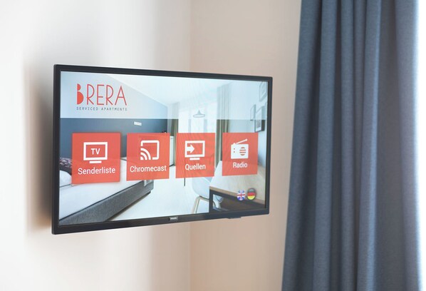 Pantalla digital en una habitación de hotel que anuncia apartamentos con servicios BRERA, con comodidades como TV, Senderliste, Chromecast, Quellen y Radio.