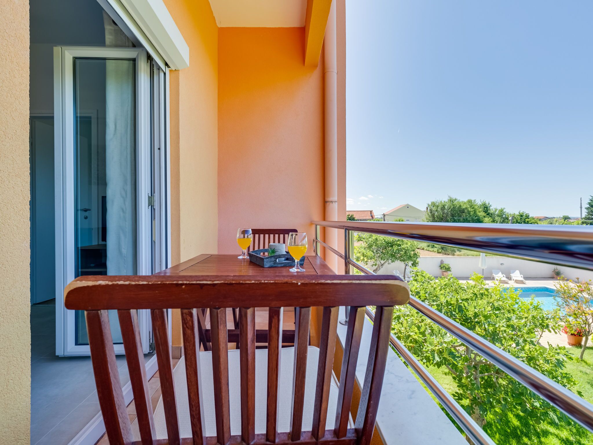 A balcony with a wooden table and chairs, a glass door, and a view of a swimming pool and greenery.