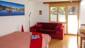 A room with a bed, a red sofa, a table with fruit, and a painting on the wall. - Sungold by Interhome (Zermatt)