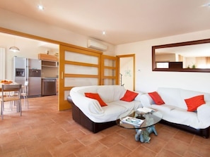 A living room with a white sofa, red cushions, a glass coffee table, and a kitchen area in the background.