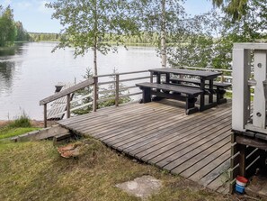 A wooden deck with picnic tables overlooking a calm lake, surrounded by trees.