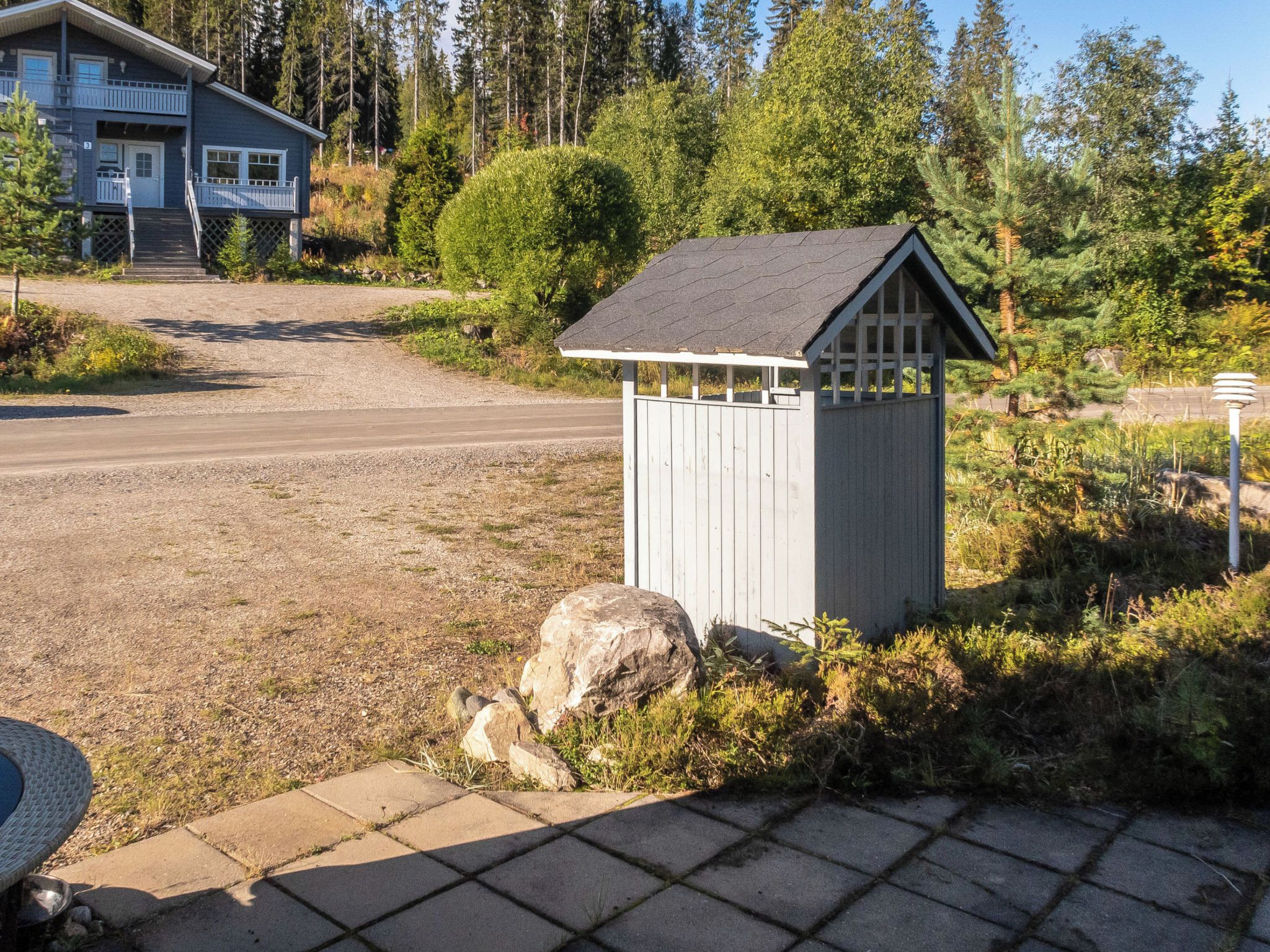 A small wooden shed with a sloped roof, situated on a gravel driveway with a large rock nearby.