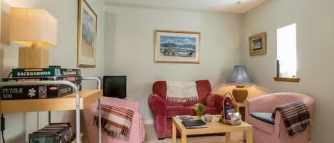 Un salon chaleureux avec un canapé rouge, un fauteuil rose, une table basse en bois et un téléviseur. On y trouve des livres et une lampe sur une étagère, ainsi qu’un tableau accroché au mur.