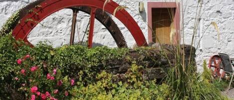 A rustic building with a large red water wheel, a wooden door, and a garden with pink flowers and green foliage.