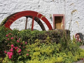 A rustic building with a large red water wheel, a wooden door, and a garden with pink flowers and green foliage. - Lorn Mill by Interhome (Balloch)