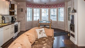 A cozy cabin interior with a wooden ceiling, a sofa with floral cushions, a dining table, and a kitchen area with a stove and coffee maker.