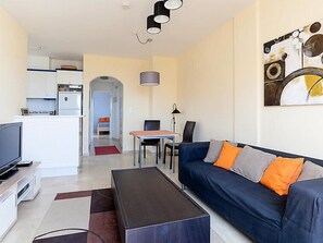 A modern living room with a sofa, coffee table, and a TV. There is a dining area with a table and chairs. A kitchen is visible in the background.