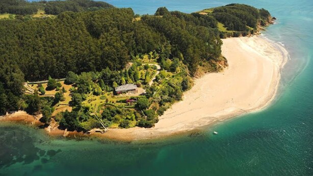 Vue aérienne d’une propriété située sur la côte, avec une plage de sable, une forêt dense et un petit bâtiment entouré de verdure.