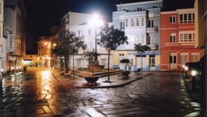 Une vue nocturne d’une rue pavée, bordée de bâtiments des deux côtés, avec une fontaine centrale et des lampadaires.