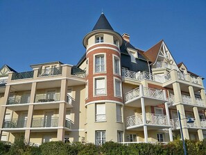 Un edificio de varios pisos con una torre distintiva que presenta un techo cónico, múltiples balcones con barandillas blancas y una fachada color beige con detalles en ladrillo rojo.