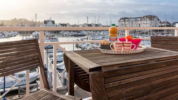 Ein Holzterrasse mit einem Frühstückstisch mit Blick auf einen Hafen voller Boote und Gebäuden im Hintergrund.
