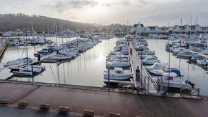 Ein Yachthafen mit zahlreichen Booten, die in ruhigem Wasser liegen, einem Fußweg und einer Kulisse aus Hügeln und Gebäuden.
