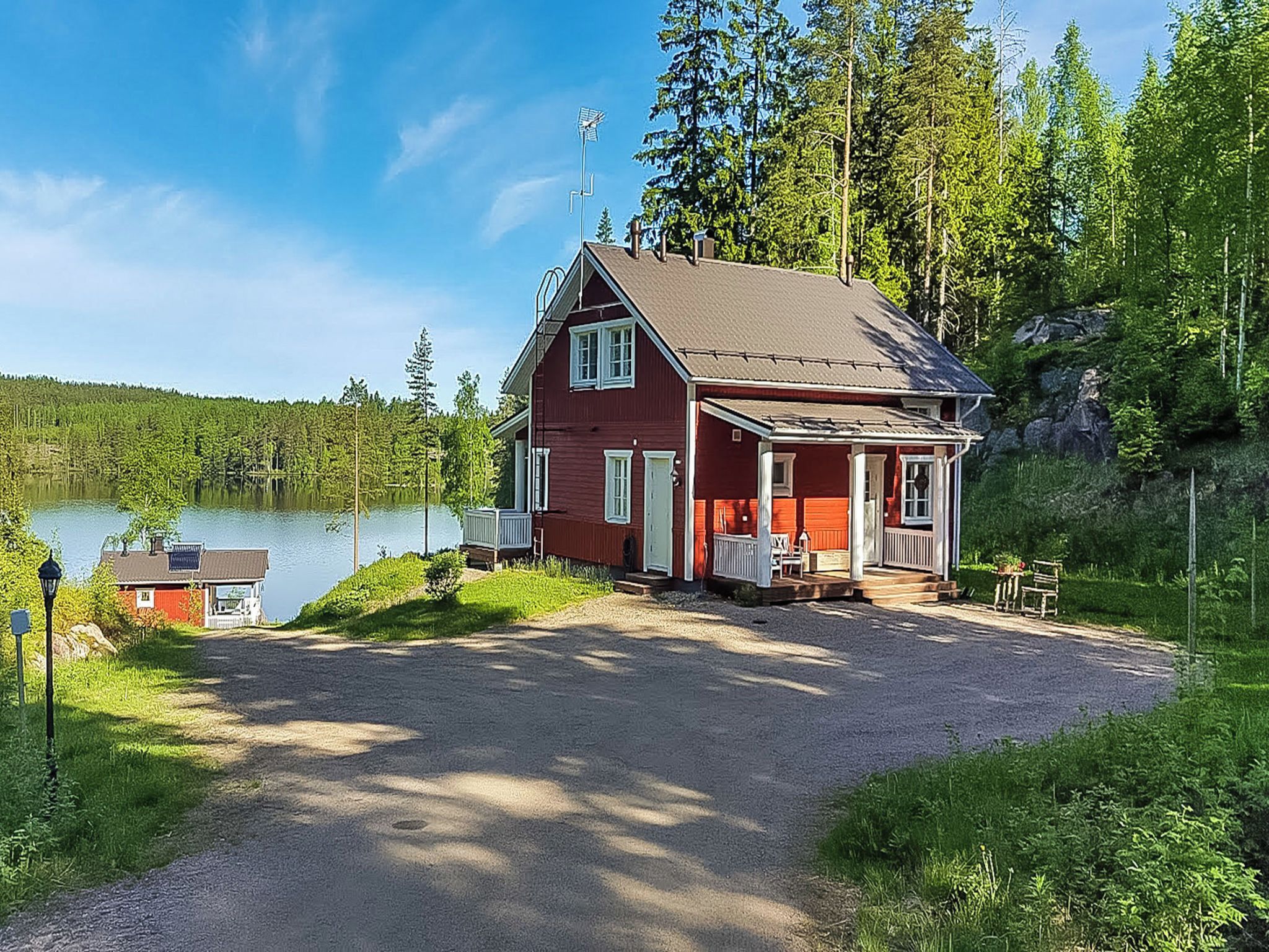 Punainen mökki, jossa on kuisti, sorapäällysteinen ajotie ja taustalla järvi.