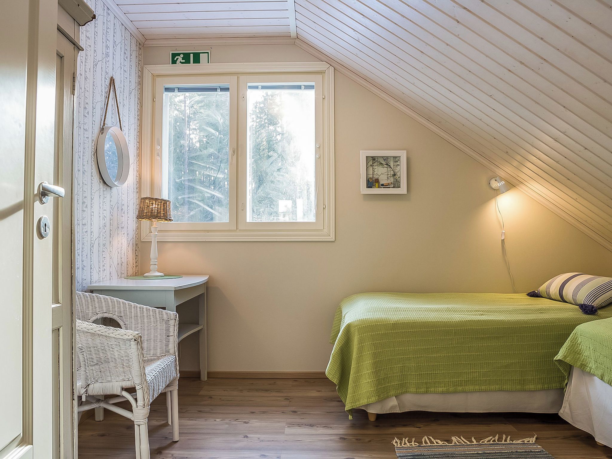 Ett rum med ett sluttande tak, ett fönster med frostat glas, ett skrivbord med en stol, en säng med grönt sänglinne och en tavla som hänger på väggen.