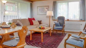 A cozy living room with a wooden ceiling, a sofa with red and white cushions, a blue armchair, a wooden coffee table, and a patterned rug. - Zur Linde Apt.W by Interhome (Wengen)