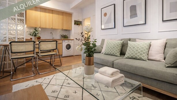 A modern living room with a sofa, a coffee table, and a dining area with chairs. There is a kitchen in the background with wooden cabinets and a washing machine.