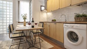 A modern kitchen with wooden cabinets, a built-in washing machine, a dining table with chairs, and a window with a view of trees.