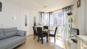 Una sala de estar con un sofá gris, una mesa de comedor con sillas, un armario blanco y una ventana con cortinas.