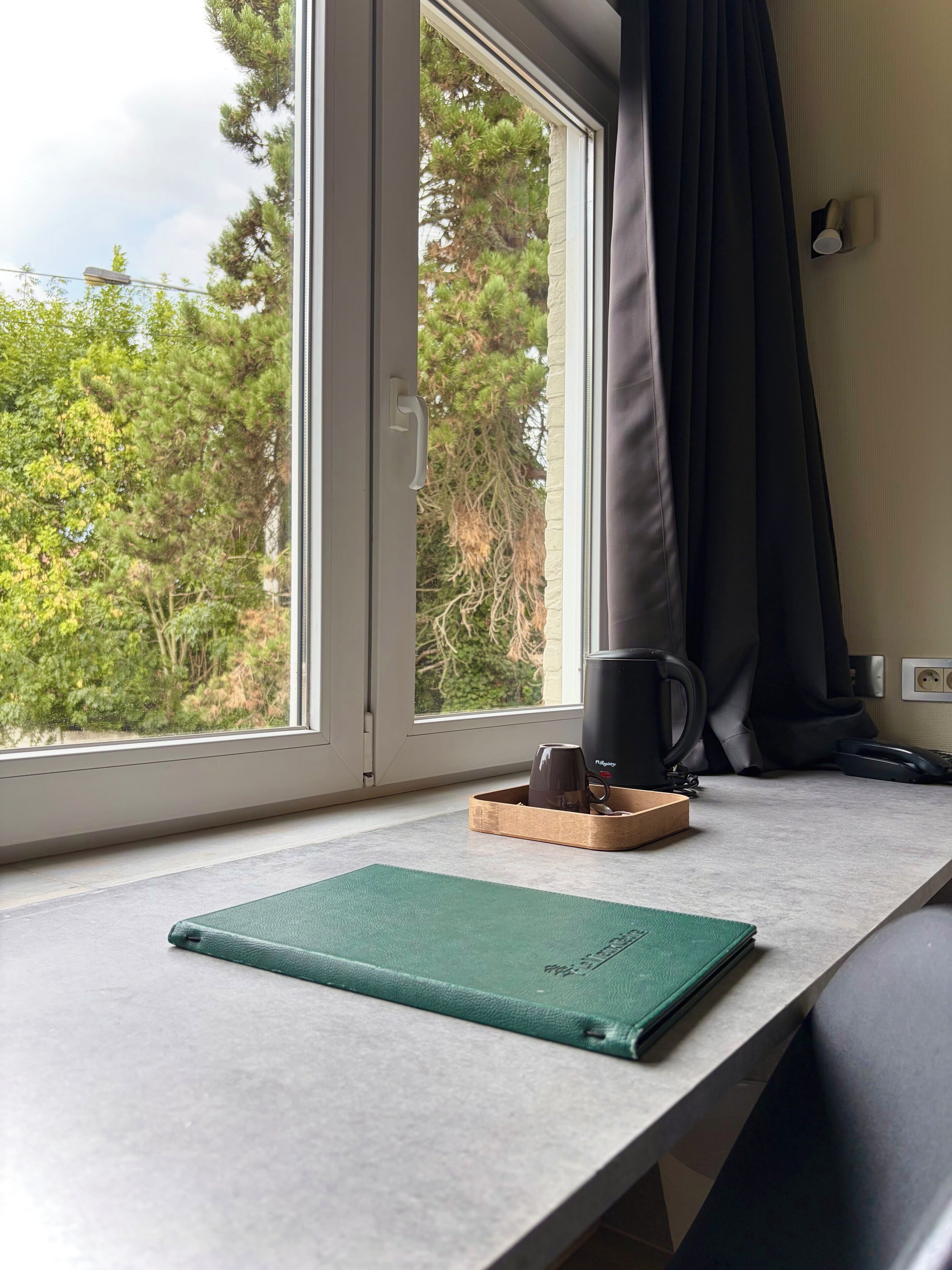 Une pièce avec une fenêtre, une table, un carnet vert, une tasse et une bouilloire.