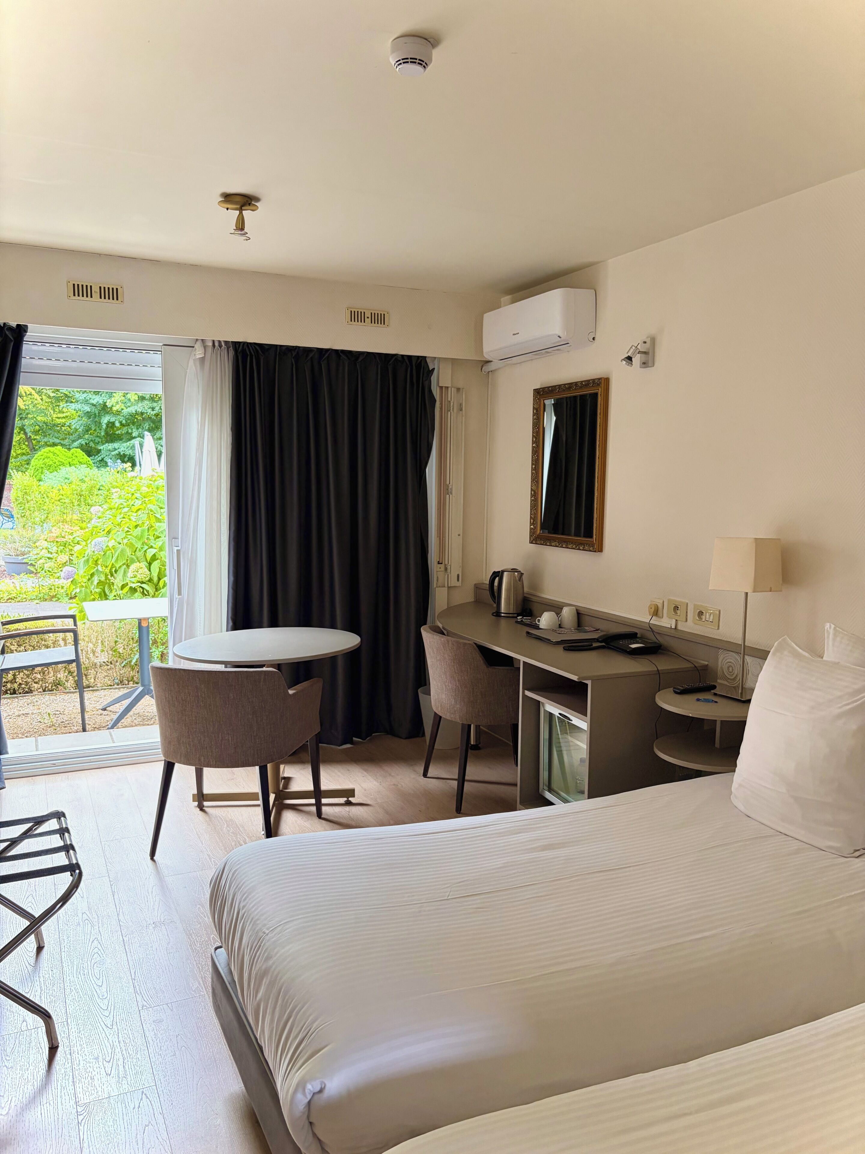 A hotel room with a bed, a desk, a chair, a small table, a coffee maker, a lamp, and a window with curtains.