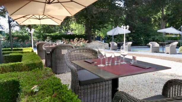 Een terras buiten met rieten meubels en tafels, omgeven door netjes bijgesnoeide hagen en bomen.