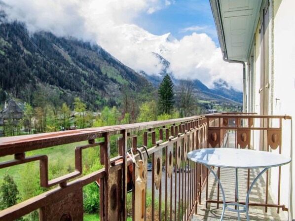 A balcony with a metal railing, a table, and chairs, overlooking a mountainous landscape with a village below.