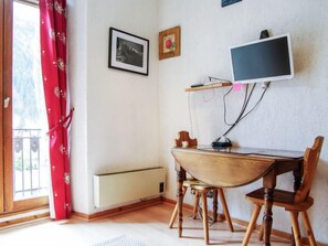 A room with a wooden table, two chairs, a TV mounted on the wall, a radiator, and a window with red curtains.