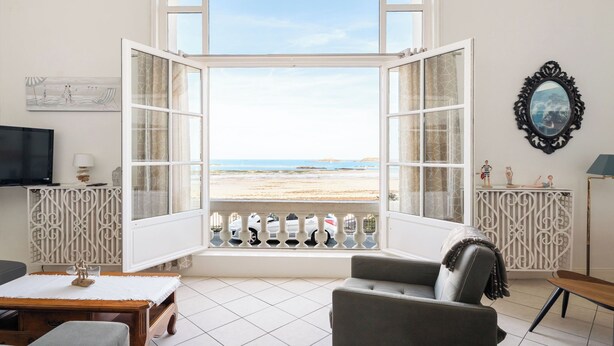 A room with a large arched window overlooking a beach, a television, a sofa, and decorative items. - La Plage by Interhome (Dinard)