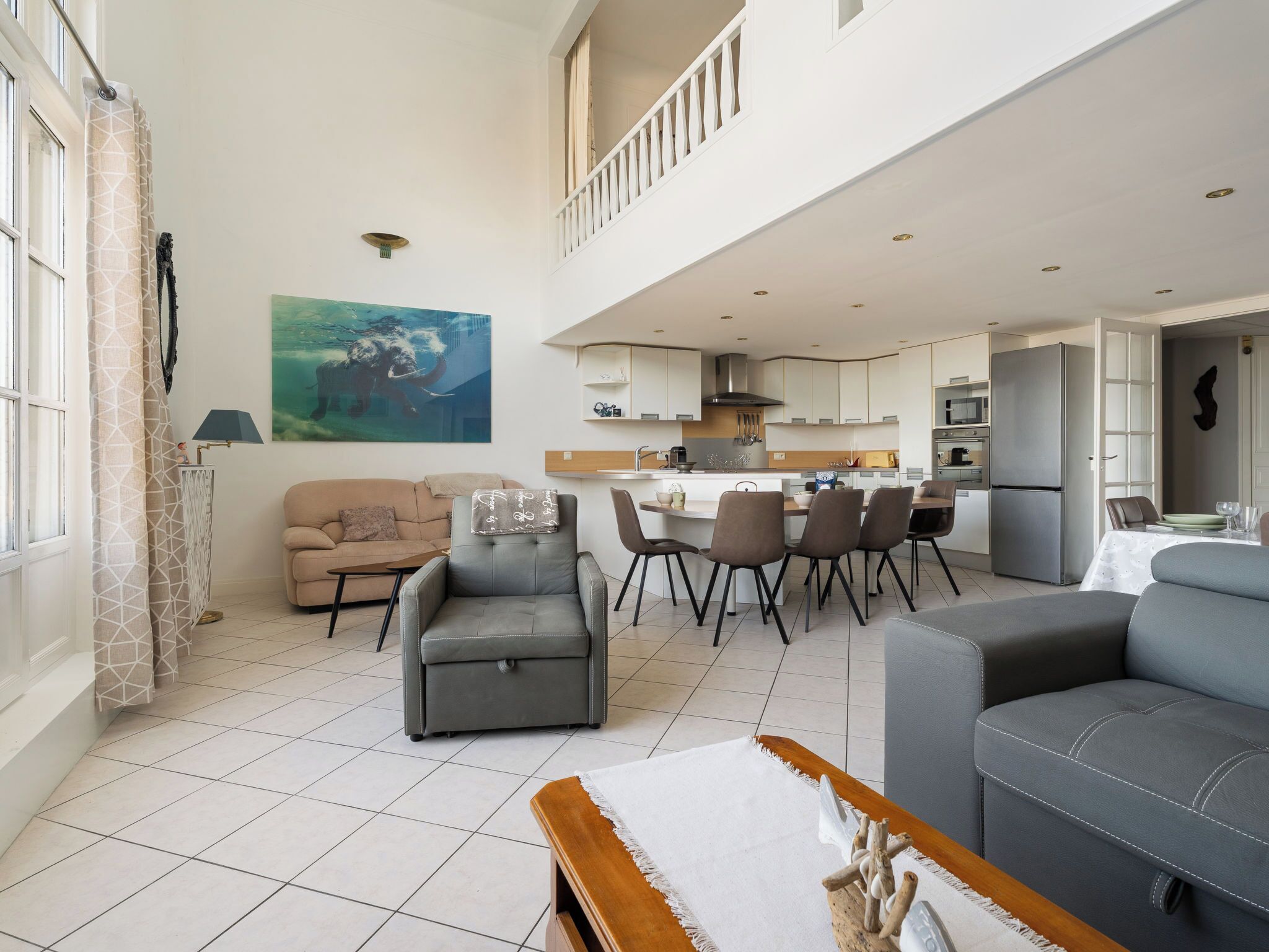 A spacious living area with a high ceiling, a dining area with a kitchen in the background, and a large window on the left.