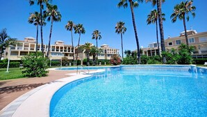 Una piscina con palmeras, cielo azul despejado y un edificio con balcones al fondo.