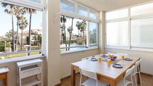 Un comedor con una mesa dispuesta para el desayuno, ventanas amplias que ofrecen vistas a palmeras y una piscina, y una silla blanca.