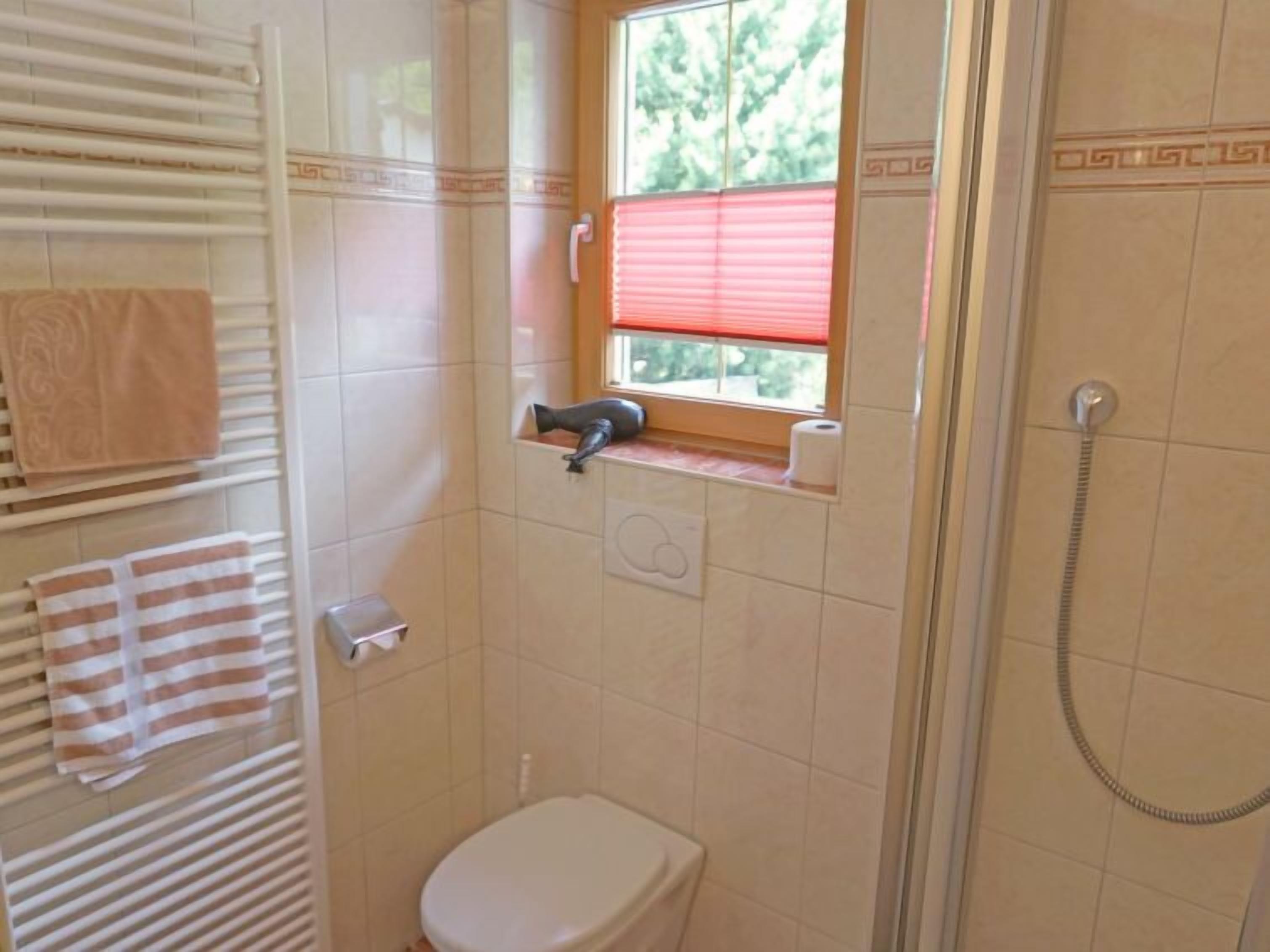 Um banheiro pequeno com vaso sanitário, porta-toalhas, janela com cortina vermelha e área de chuveiro.