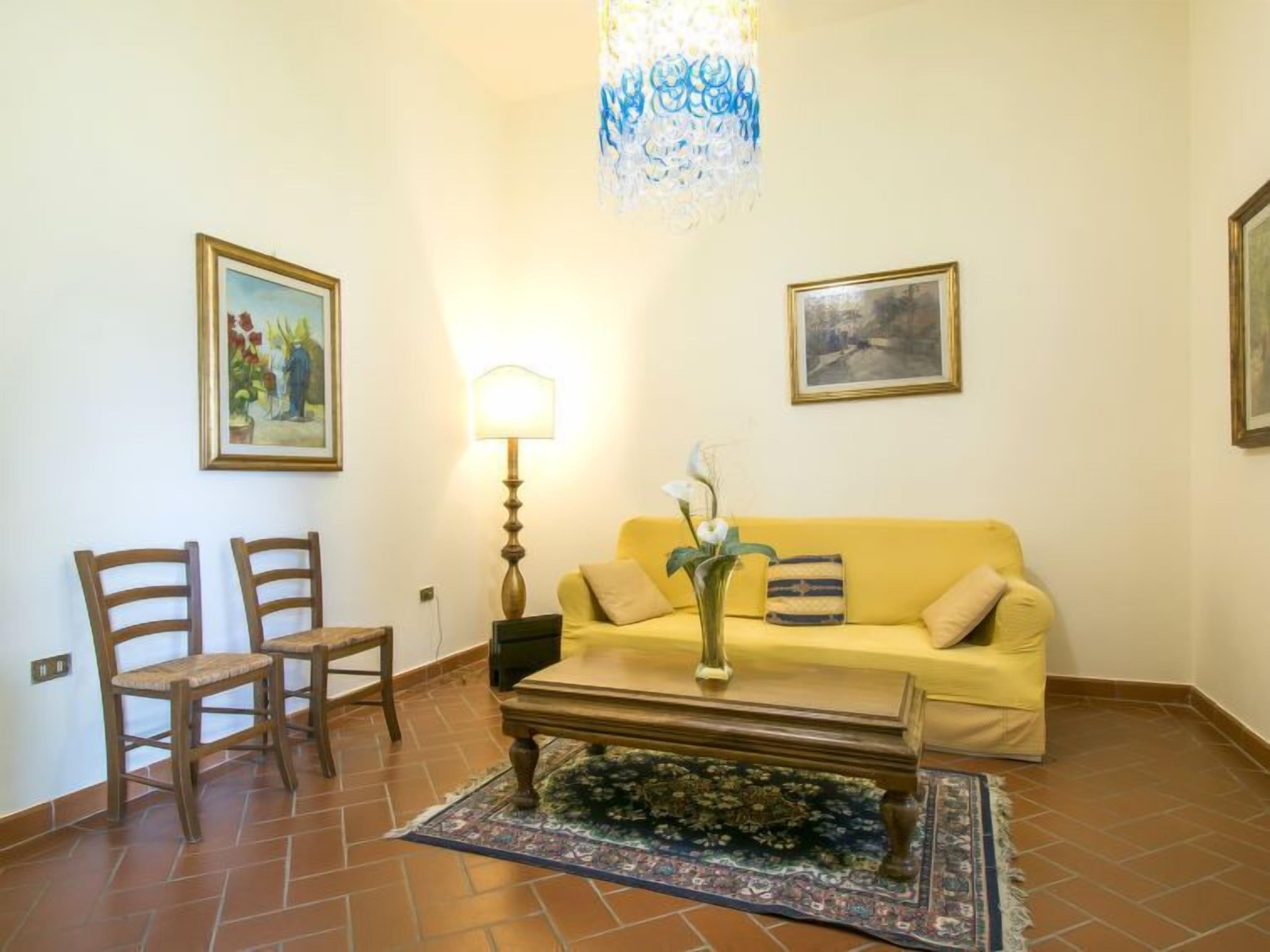 Un salon avec un canapé jaune, deux fauteuils en bois, une table basse, une lampe et deux tableaux encadrés accrochés au mur.