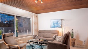 Un salon moderne avec un plafond en bois, une grande fenêtre offrant une belle vue, un canapé marron, un fauteuil en bois et une table basse.