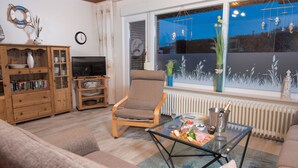 Un salon avec un canapé, une table basse en verre sur laquelle se trouvent des aliments, un meuble en bois, un téléviseur et une fenêtre donnant sur le ciel et les plantes.