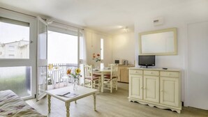 A bright, well-lit room with a bed, a dining table with chairs, a small kitchen area with a TV, and a balcony with a view of buildings. - Les Amandiers by Interhome (Canet-Plage)
