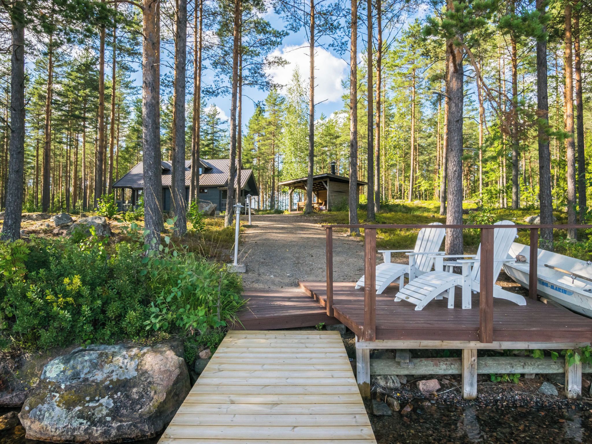 A wooden deck leading to a cabin in a forested area with a boat and lounge chairs.