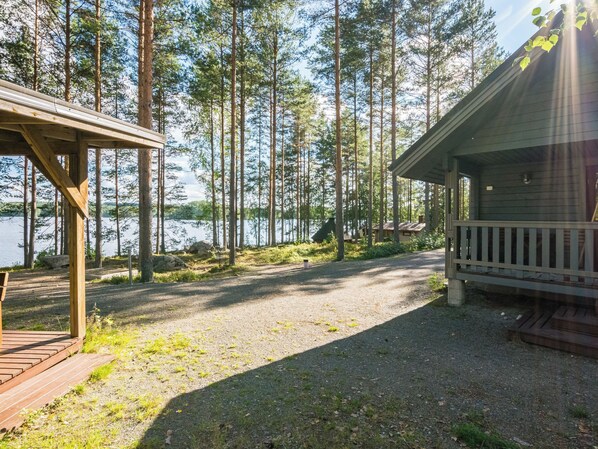 En hytte med terrasse og veranda, omgivet af høje træer og et vandareal.