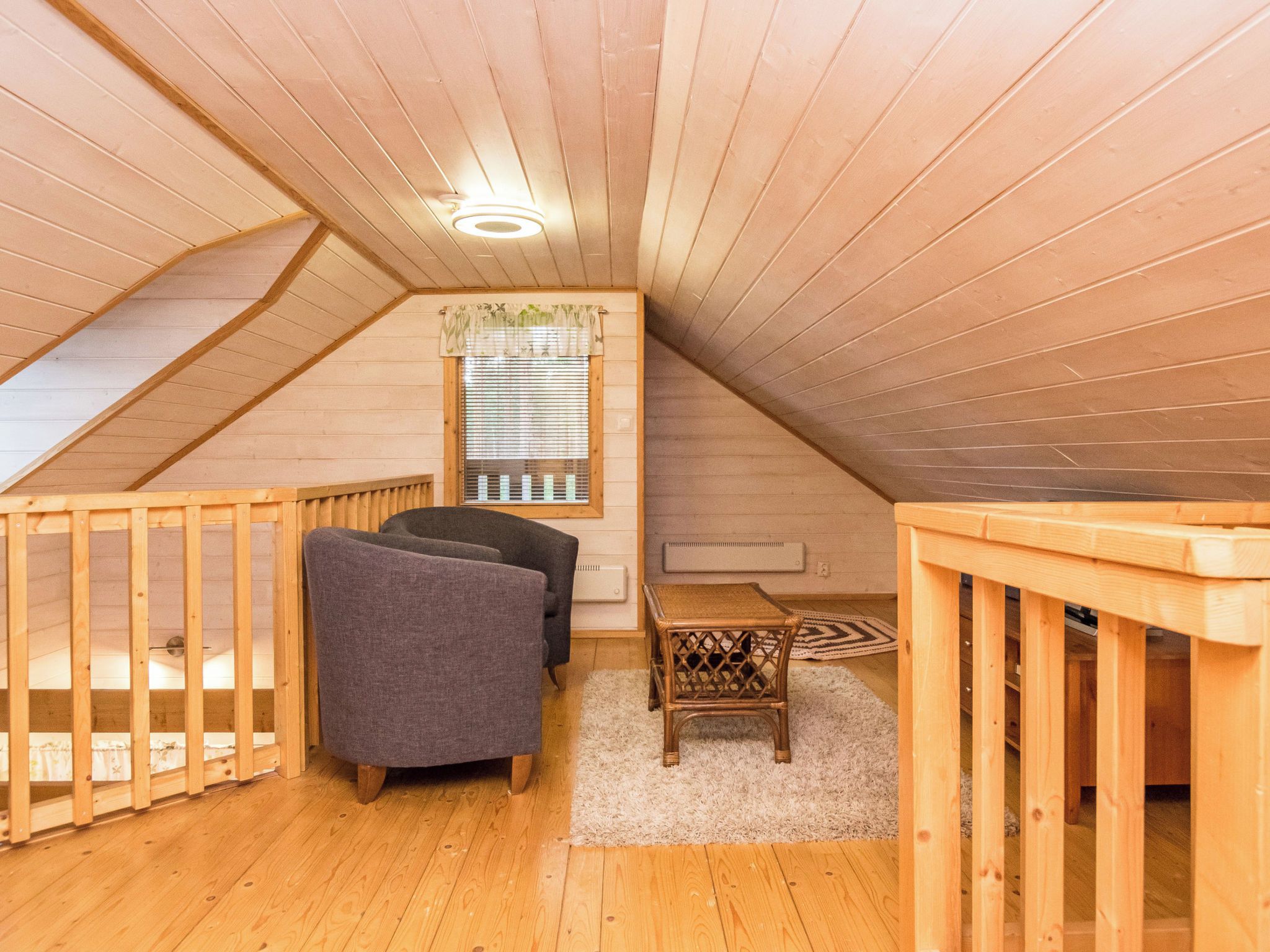 A cozy attic bedroom with a sloped ceiling, wooden walls, a window with blinds, a small table, two chairs, and a bed.
