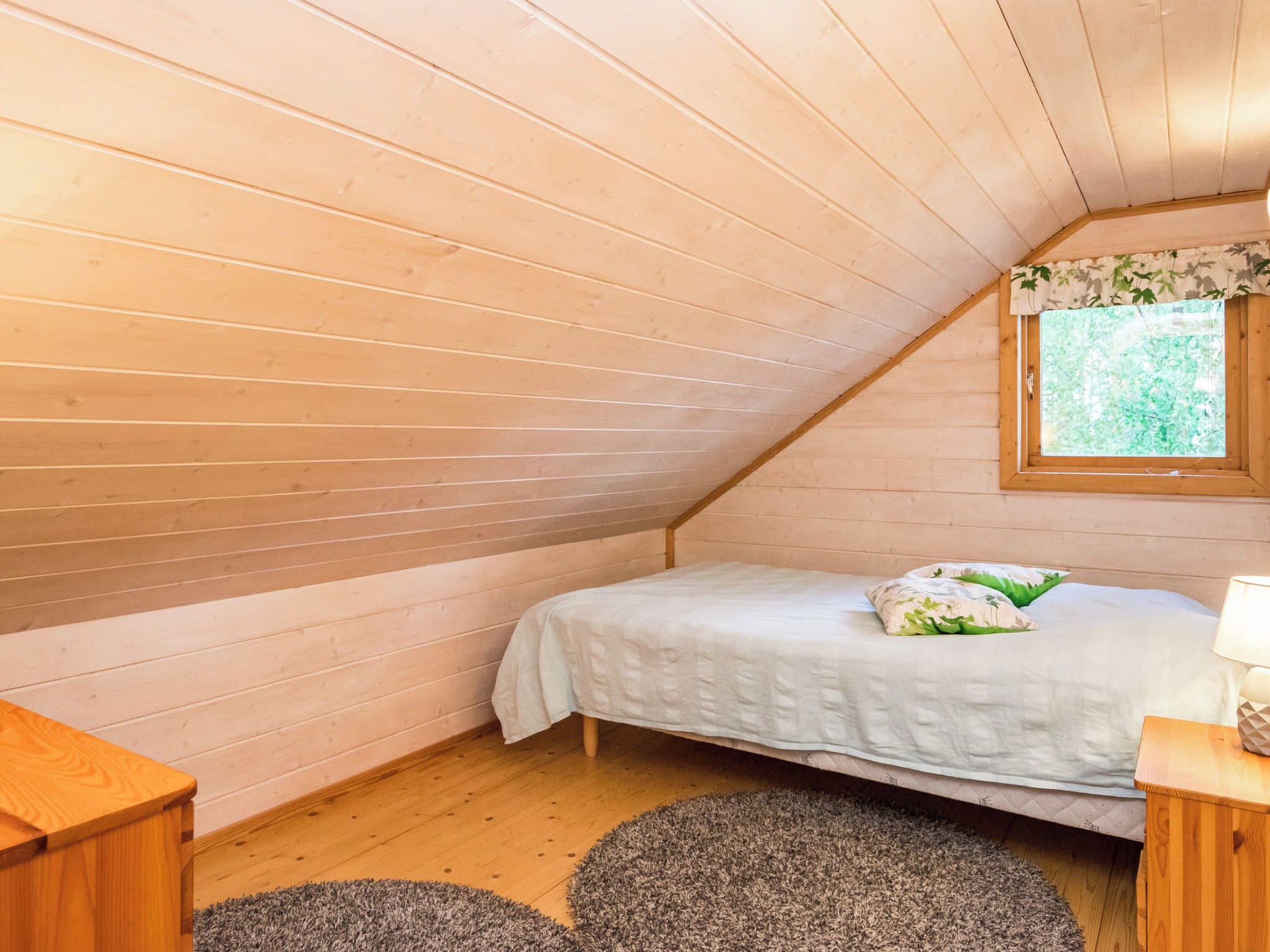 A small, cozy room with wooden walls and a sloped ceiling. There is a bed with a white bedspread and pillows, a small wooden nightstand with a lamp, and a window with a view of greenery outside.