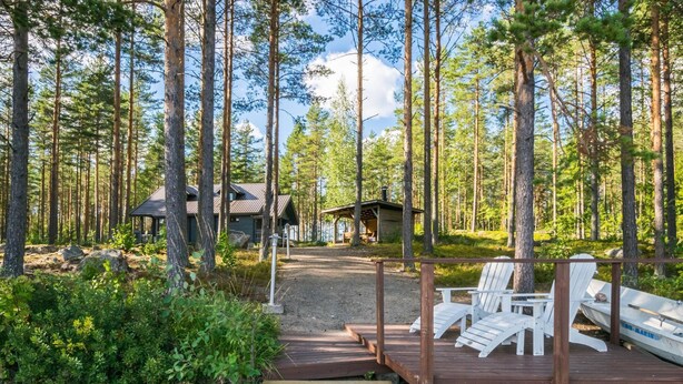 Puinen terassi, joka johtaa mökille metsäisellä alueella. Terassilla on Adirondack-tyylisiä puutarhatuoleja ja lähellä on vene.