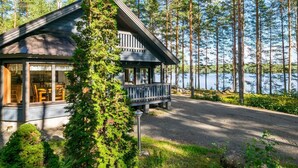 Mökki, jossa on kuisti ja suuret ikkunat, joista avautuu näkymä järvelle. Mökkiä ympäröivät korkeat puut.