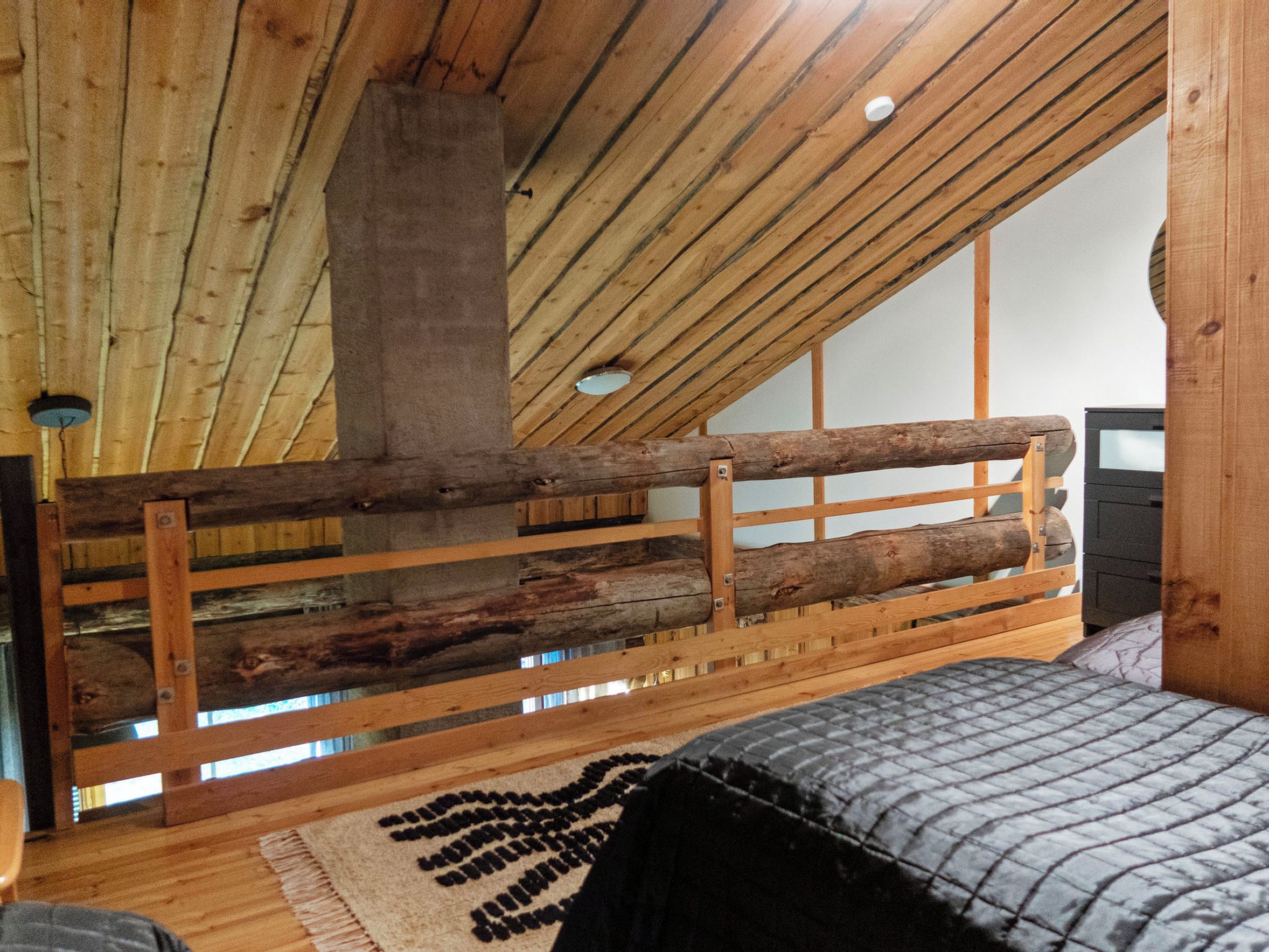 Une chambre avec un plafond en pente, des poutres apparentes, un lit recouvert d’une couette grise et une tête de lit en bois.